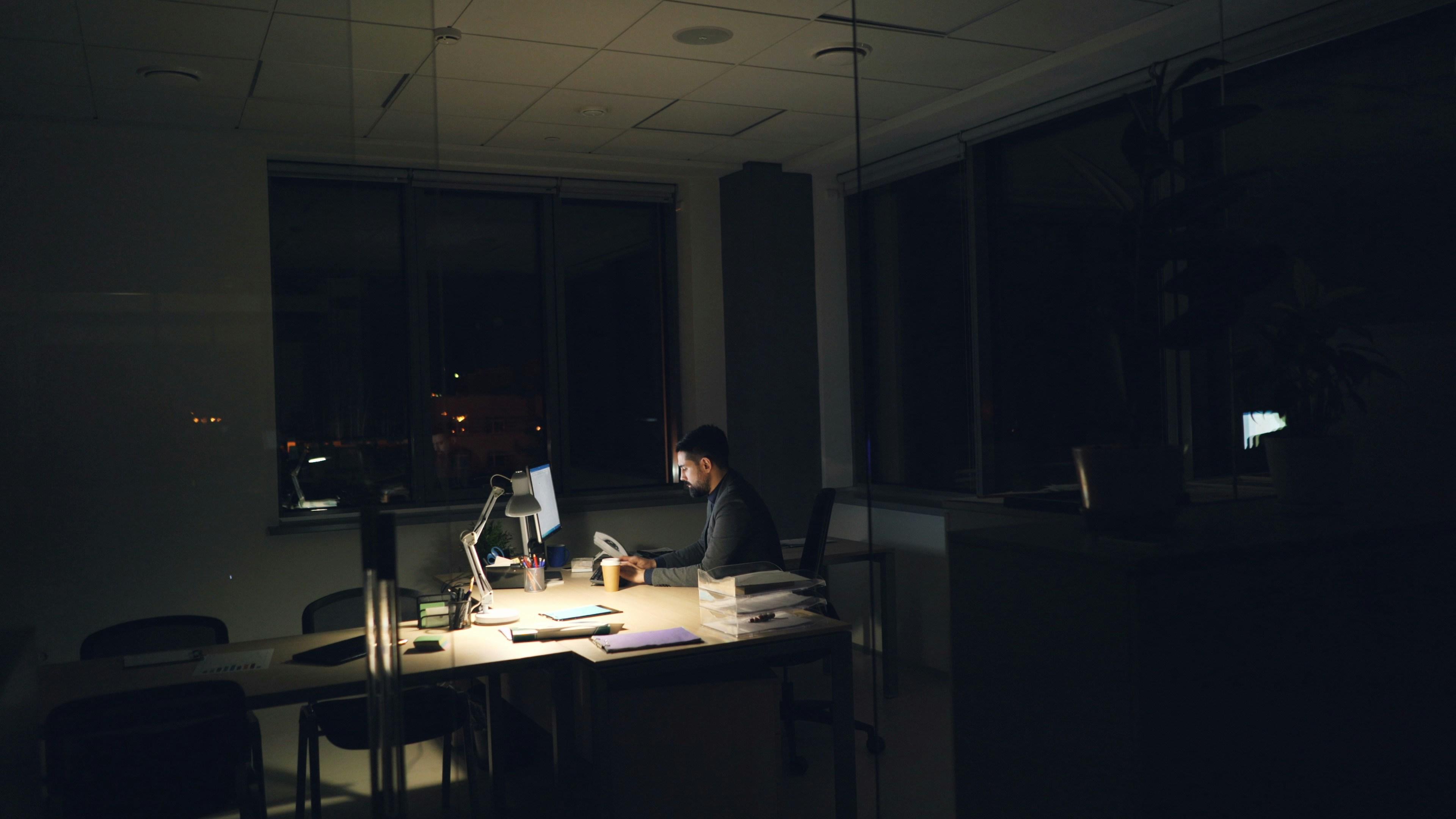 A lone figure working late at a lamp-lit desk in an otherwise dark office at night.