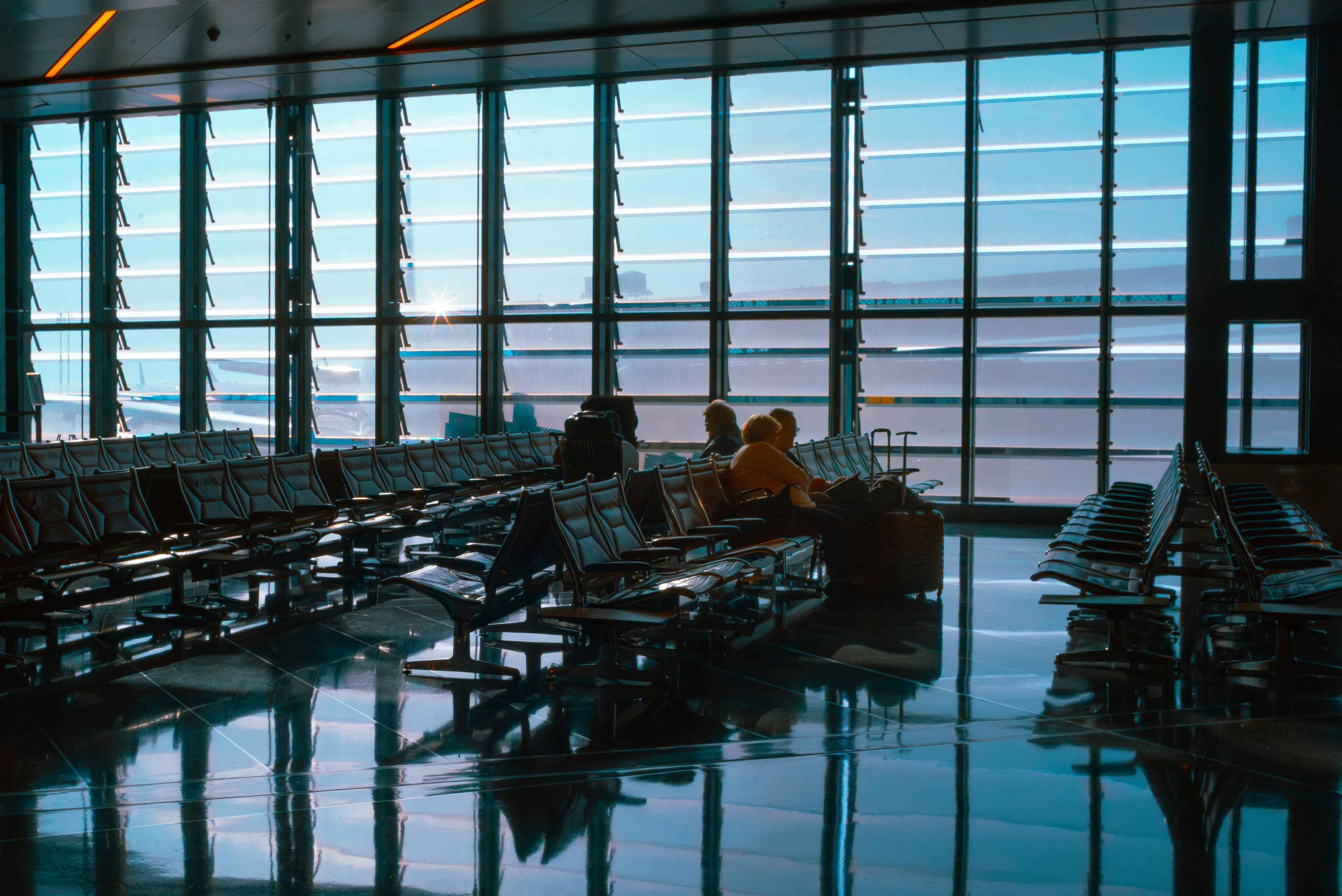 Silhouetted travelers seated near a large terminal window at dusk, reflections across the floor.